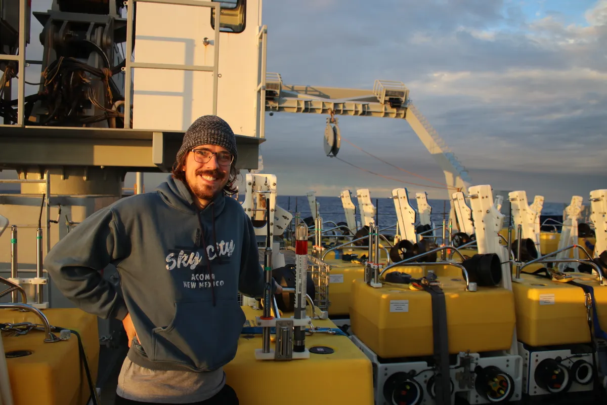 Joey Phillips on a research vessel deck beside orange ocean-bottom seismometer packages.