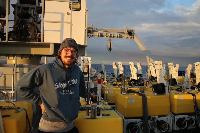 Joey Phillips on a research vessel deck beside orange ocean-bottom seismometer packages.