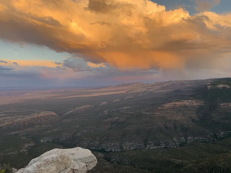 Golden sunset storm clouds trailing rain over forested ridges and a broad valley, seen from a pale rock outcrop.