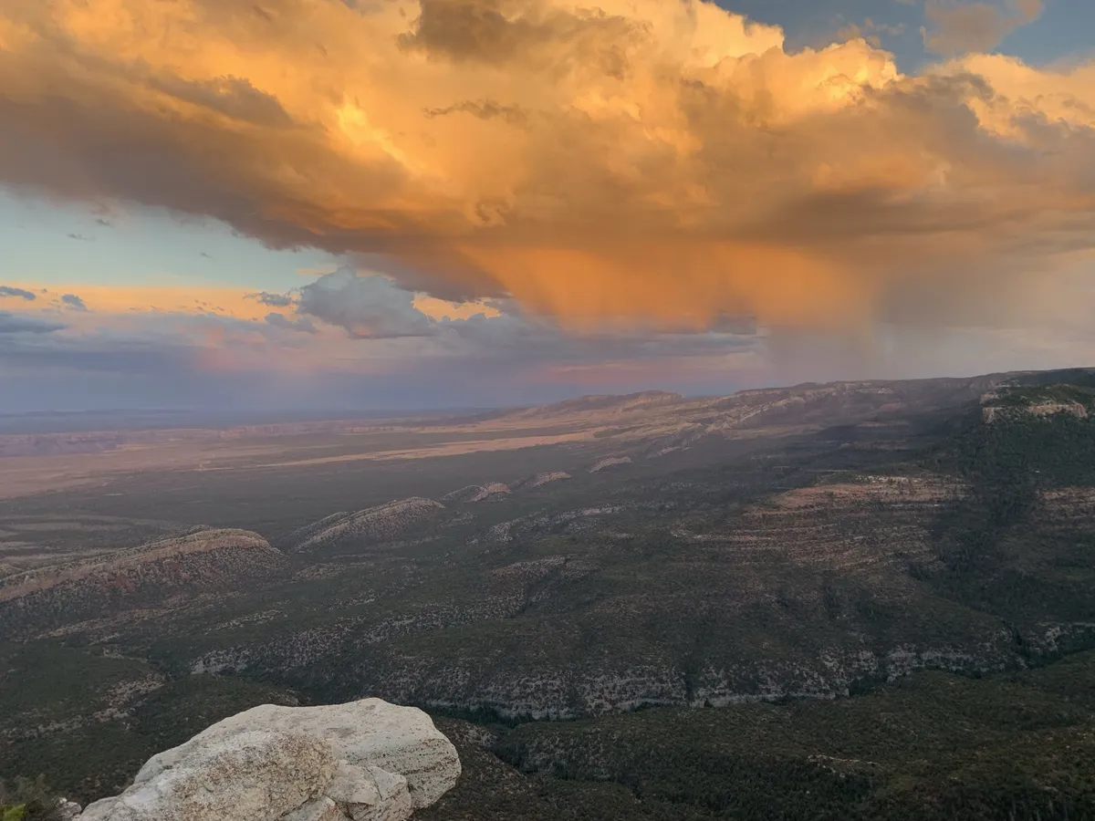 Golden sunset storm clouds trailing rain over forested ridges and a broad valley, seen from a pale rock outcrop.